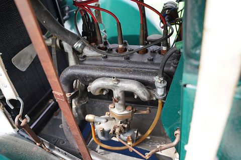 Close view of a small four-cylinder engine with copper pipes and a carburettor inside a green bonnet. Opens a larger view