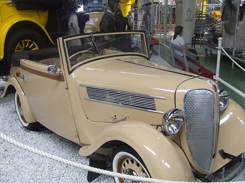 A tan open two-door car with a curved grille, faired headlamps and white-wall tyres behind a museum rope. Opens a larger view
