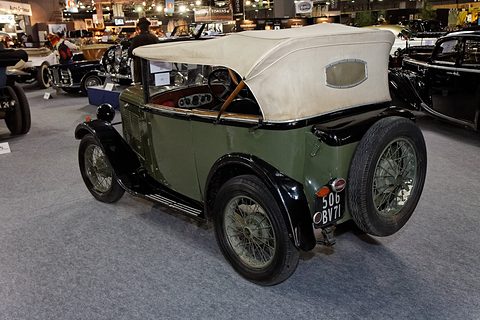 The rear of a green open tourer with a pale fabric hood and a spare wheel, on a show floor. Opens a larger view