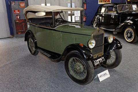 A green open four-seat tourer with black wings, wire wheels and a pale hood, among other cars on a show floor. Opens a larger view