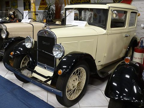 A cream four-door saloon with black wings and wire wheels stands among other old cars in a museum hall. Opens a larger view