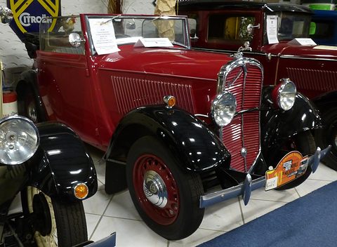 A red open car with black wings, a chrome radiator shell and red wheels stands in a crowded museum hall. Opens a larger view