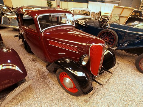 A red rounded two-door saloon with black wings and red wheels stands among other vintage cars on gravel.