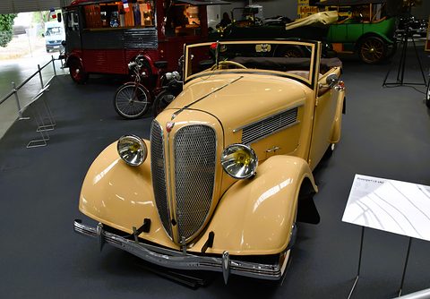 A tan open two-door car with a split grille and faired headlamps on a dark museum floor beside a display sign. Opens a larger view