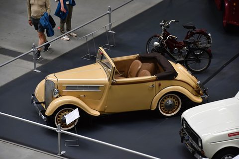 A tan open car seen from above and behind on a dark museum floor, its brown seats visible and a motorcycle beyond. Opens a larger view