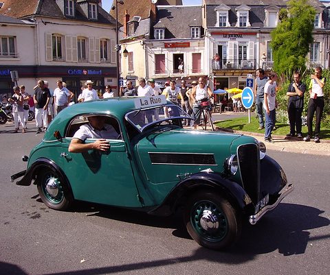 A green rounded two-door car drives round a town roundabout past spectators and old buildings. Opens a larger view