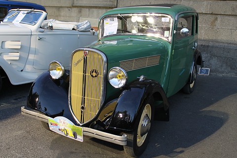 A green two-door saloon with black wings, a split grille and yellow headlamps parked on tarmac beside a white car. Opens a larger view