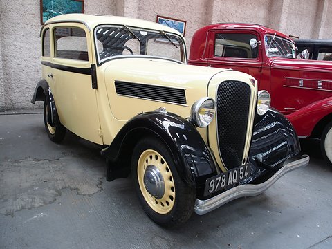 A cream two-door saloon with black wings, a black split grille and yellow headlamps indoors beside a red car. Opens a larger view