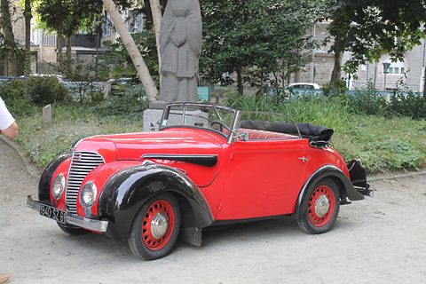 A bright red open two-door car with black wings and red wheels parked on gravel under trees by a statue. Opens a larger view