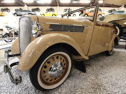 A tan open two-door car with a chrome grille, louvred bonnet and white-wall tyres on a gravel museum floor. Opens a larger view
