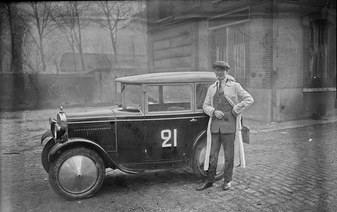 A small dark saloon numbered 21 on its door stands on cobbles beside a man in a long pale coat.