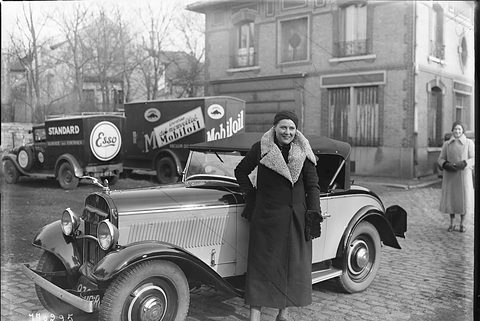 A woman in a fur-collared coat standing before a small two-door cabriolet with its hood up, Esso and Mobiloil vans behind. Opens a larger view
