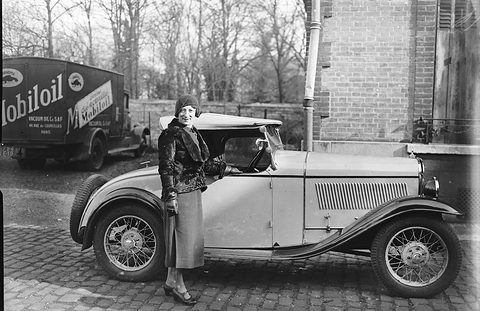 A woman in a fur jacket standing at the door of a pale two-seat cabriolet with its hood folded, a Mobiloil van behind. Opens a larger view