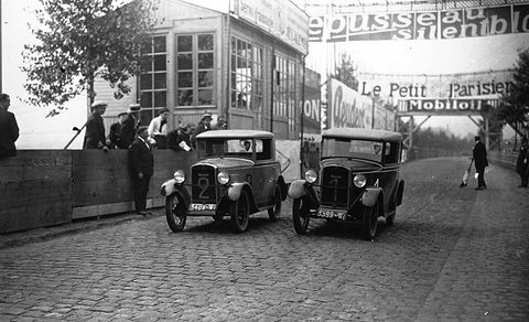 Two small square-roofed saloons side by side on a wide cobbled road, spectators behind a fence and advertising banners overhead.