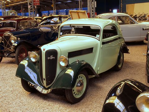 A pale green rounded coupé with dark green wings and a split grille stands among other vintage cars on gravel. Opens a larger view