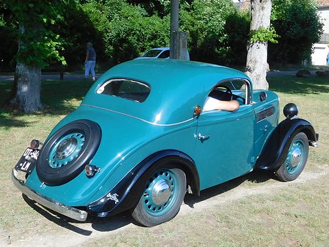 A turquoise two-door coupé with black wings and a spare wheel set into its sloping tail, parked on grass under trees. Opens a larger view
