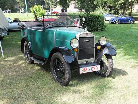 A small turquoise open tourer with black wings and wire wheels parked on grass, its hood folded. Opens a larger view