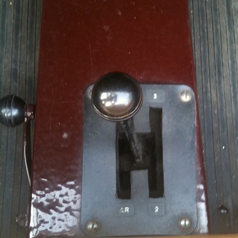 Close view of a chrome-knobbed gear lever in an H-shaped gate on a maroon floor panel. Opens a larger view