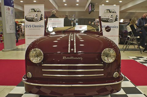 Front view of a maroon open two-seater with round headlamps, chrome bonnet strips and grille bars. Opens a larger view