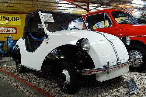 White doorless two-seater with black cycle-style wings and a black hood among museum cars. Opens a larger view