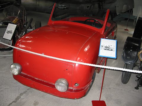 Red doorless two-seater with a rounded nose and two headlamps behind a white rope. Opens a larger view