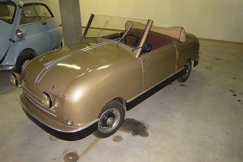 Gold-coloured open two-seater with a folded hood in a plain garage beside a pale blue car. Opens a larger view