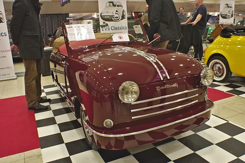 Maroon open two-seater on a chequered floor beside a dealer’s banner, visitors behind.