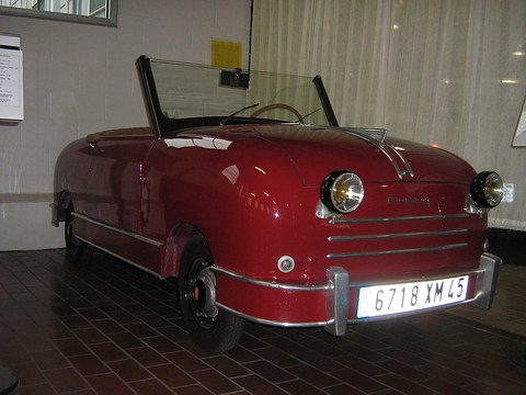 Maroon open two-seater with a slatted grille, round headlamps and a chrome bumper on a brick floor. Opens a larger view