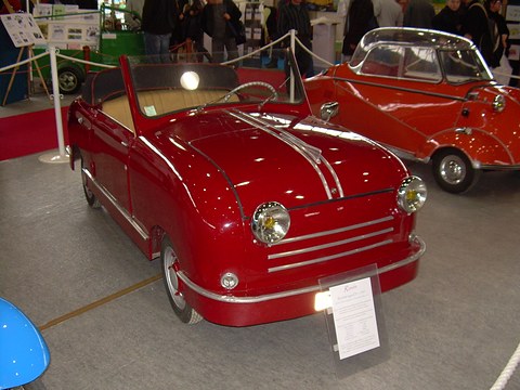 Red open two-seater with chrome bonnet strips behind a rope, a red Messerschmitt beside it. Opens a larger view