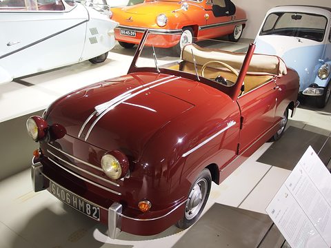 Maroon open two-seater with chrome bonnet strips and white coachlines, other microcars around it. Opens a larger view