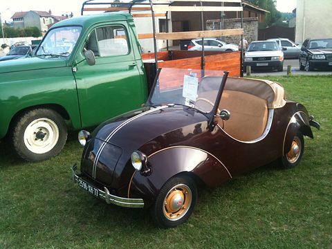 Brown doorless two-seater with cream seats and orange wheels on grass beside a green pick-up. Opens a larger view