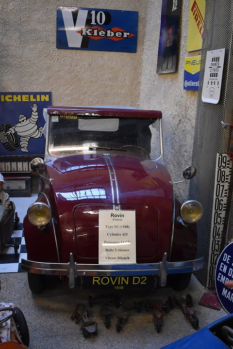 Maroon doorless two-seater with a black hood and a white information card, enamel signs behind. Opens a larger view