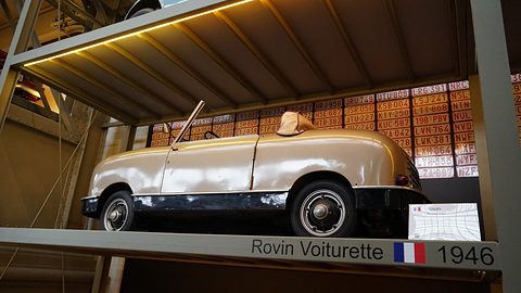 Cream doorless two-seater seen from below on a lit shelf, a wall of number plates behind. Opens a larger view