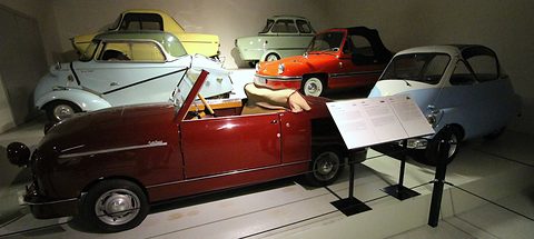Maroon open two-seater beside an information stand, bubble cars and small coupés behind it.