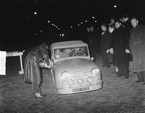 A small rounded French two-seater with rally plate 204 under night lights, two bystanders at the right.
