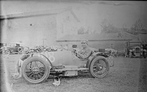 Black-and-white: a man in goggles sits in a stark open racing car with wire wheels, parked on grass before a farm building. Opens a larger view