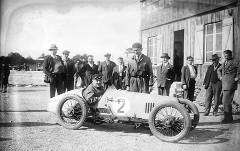 Black-and-white: a pale open racing car with a large number 2, men in caps and hats standing behind it.