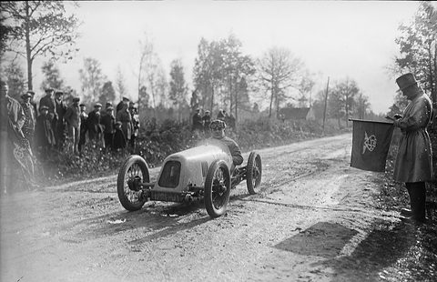 Black-and-white: a small open racing car on a muddy country road, spectators on the bank and a marshal with a flag. Opens a larger view