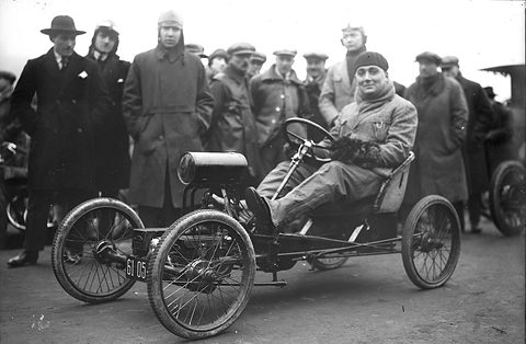 Black-and-white: a man in a cap sits on a skeletal four-wheeled chassis with wire wheels, a crowd in coats behind.