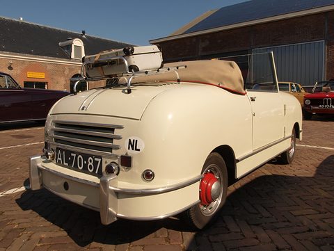 Rear of a cream open two-seater with a slatted engine cover, NL plate and luggage on the rack. Opens a larger view