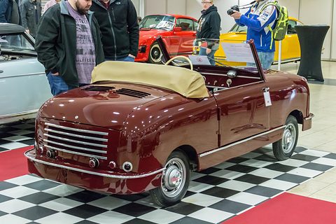 Rear of a maroon open two-seater with a slatted engine cover and folded hood on a chequered floor. Opens a larger view