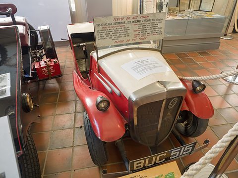 Small red and cream open car with its round-the-world board on the windscreen frame and a sheet of paper on the bonnet. Opens a larger view