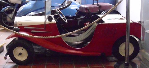 Left side of a tiny red open car with cream bonnet and white wheels, a rope and steel post in front, scooters behind.