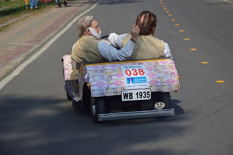 Rear of a miniature car covered in printed banknotes, plate WB 1935 and rally number 038, two occupants facing away. Opens a larger view
