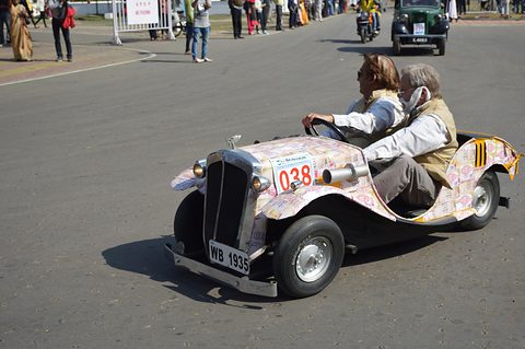 Miniature open car printed with a pattern of banknotes, two older passengers aboard, rally road behind. Opens a larger view