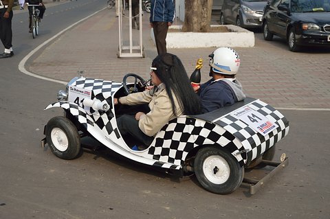 Black and white chequered miniature car turning on a road with a driver and a helmeted passenger aboard. Opens a larger view