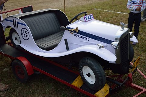 White miniature open car marked AC TAXI and NO REFUSAL, with a roof-style taxi sign, standing on a red trailer. Opens a larger view