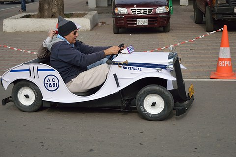 Side view of a white miniature open car with a capped driver whose knees rise above the body, a traffic cone ahead. Opens a larger view