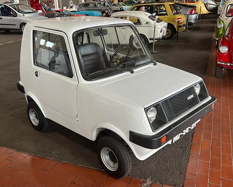 Small white two-door city car with a black grille panel and black bumpers on a brick floor among other small cars.
