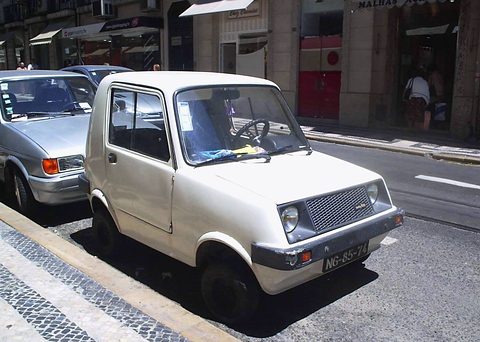 Small white two-door car with a black grille parked in sunlight beside a mosaic pavement, a silver saloon behind.